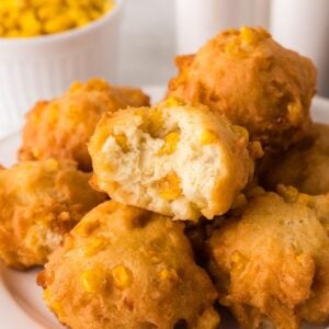 A plate of golden, fluffy corn fritters with visible corn kernels. One fritter is broken open, revealing a soft, airy interior. A bowl of corn kernels is visible in the background.