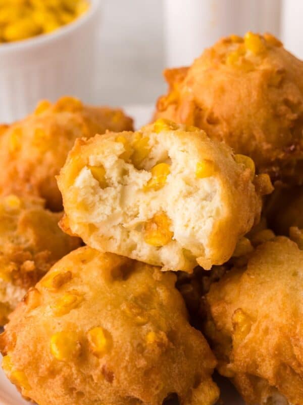 A plate of golden, fluffy corn fritters with visible corn kernels. One fritter is broken open, revealing a soft, airy interior. A bowl of corn kernels is visible in the background.