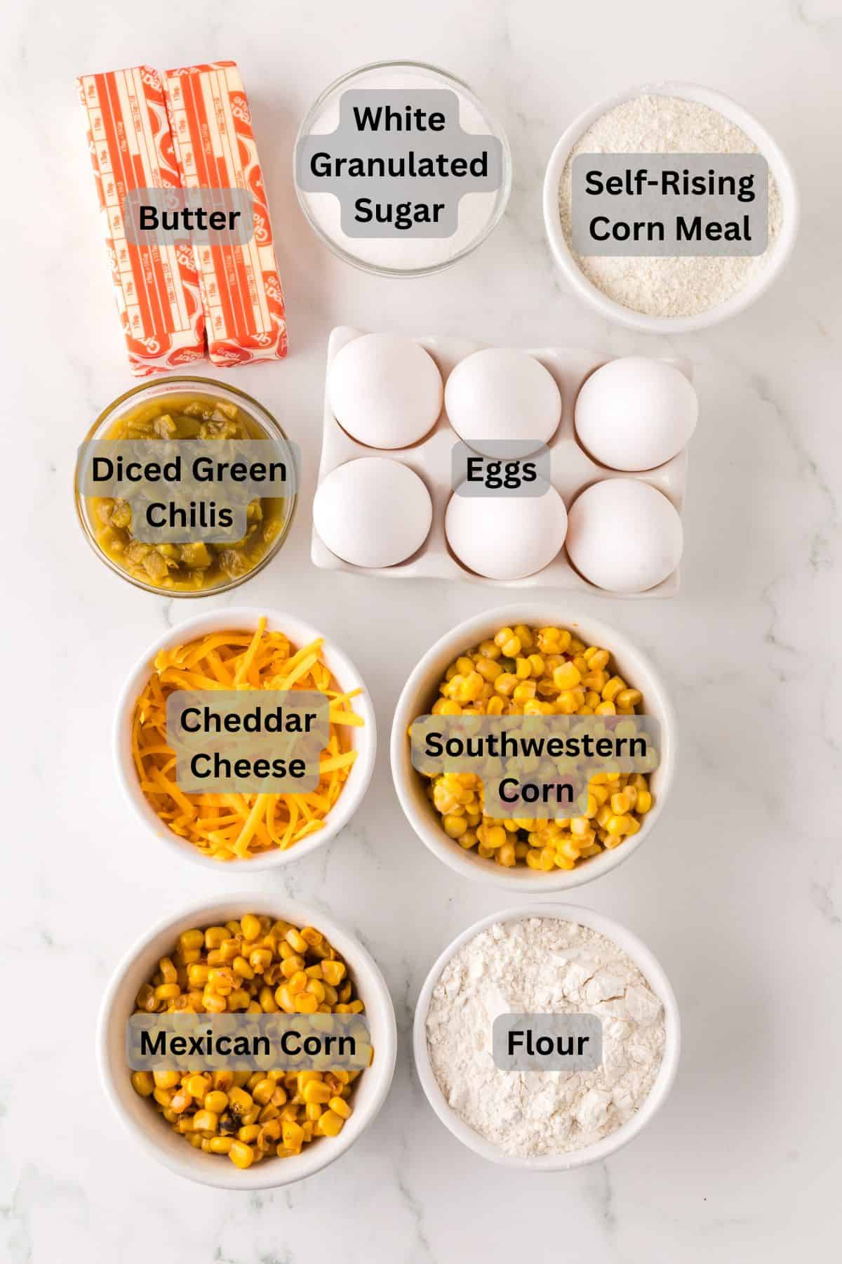 Ingredients for a recipe on a marble surface, including butter, white granulated sugar, self-rising corn meal, diced green chilis, eggs, cheddar cheese, southwestern corn, Mexican corn, and flour, each labeled in bowls or packaging.