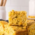 mexican cornbread recipe featuring a stack of golden cornbread squares with visible corn kernels, set against a light kitchen background. A jar with wooden utensils is blurred in the back. The focus is on the texture of the top piece.