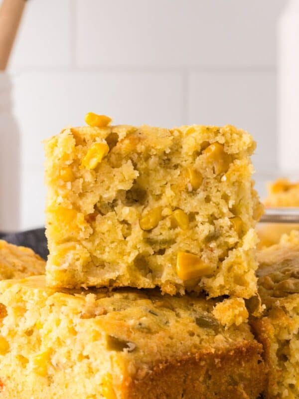 mexican cornbread recipe featuring a stack of golden cornbread squares with visible corn kernels, set against a light kitchen background. A jar with wooden utensils is blurred in the back. The focus is on the texture of the top piece.