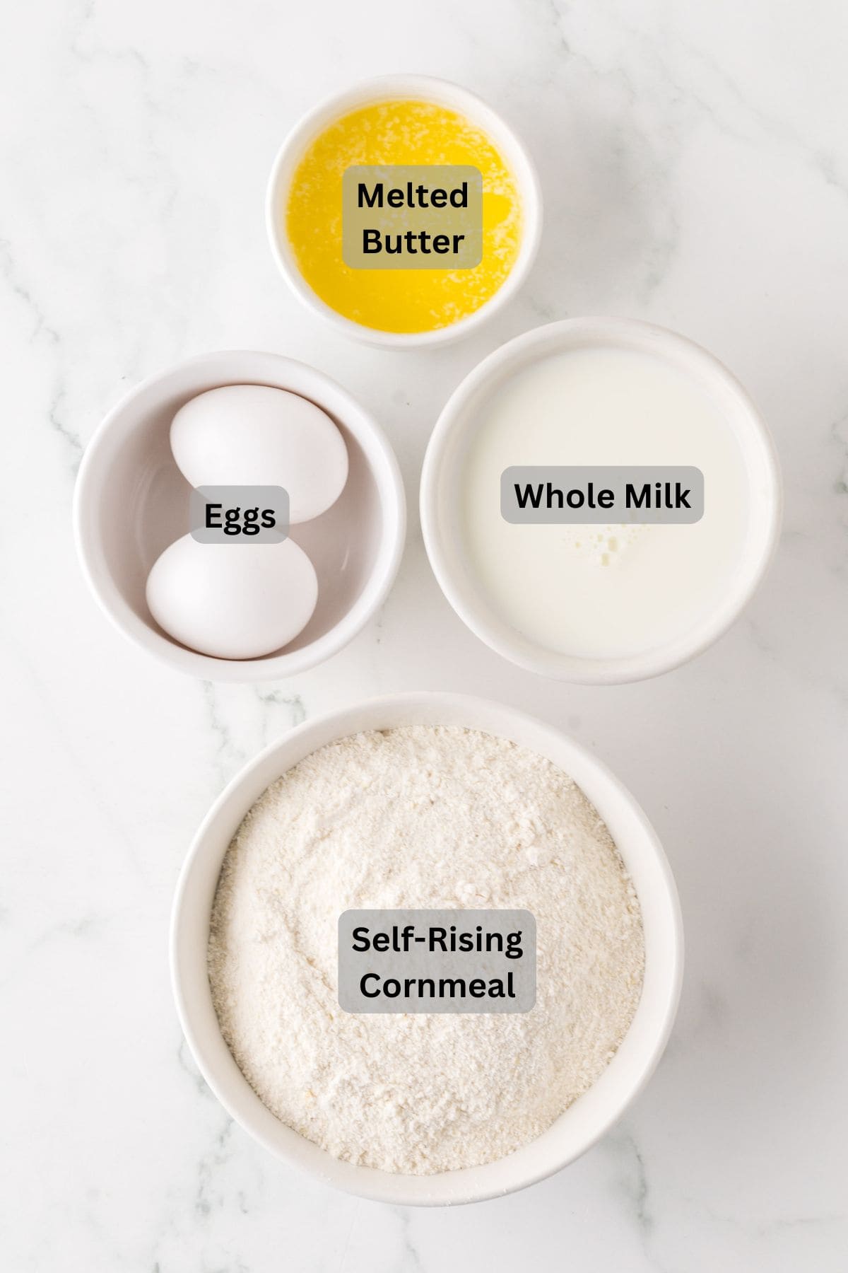 Top-down view of labeled baking ingredients on a marble surface: a bowl of melted butter, a bowl of whole milk, two eggs in a bowl, and a bowl of self-rising cornmeal.