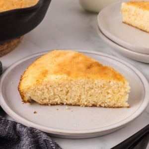 A slice of golden Southern cornbread on a gray plate. The texture looks moist and crumbly. In the background, theres a partial view of another slice and a skillet with more cornbread. A dark cloth napkin is on the left, enhancing this homestyle recipes rustic charm.