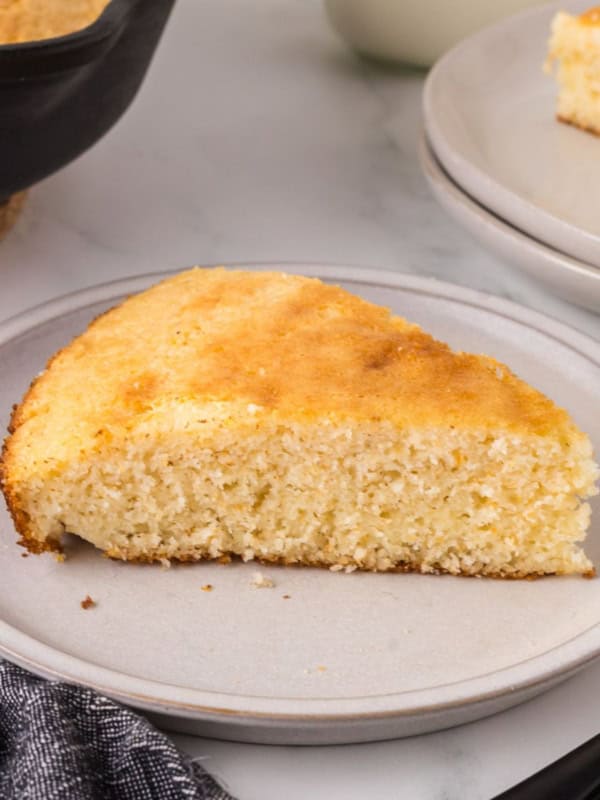 A slice of golden Southern cornbread on a gray plate. The texture looks moist and crumbly. In the background, theres a partial view of another slice and a skillet with more cornbread. A dark cloth napkin is on the left, enhancing this homestyle recipes rustic charm.