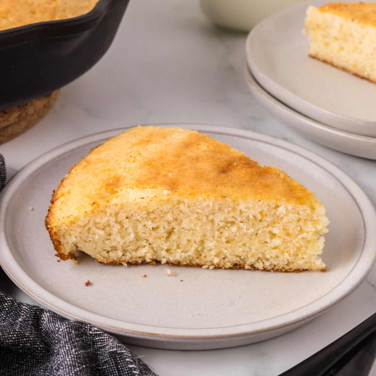 A slice of golden cornbread on a round white plate. The texture appears moist and fluffy. In the background, part of a skillet with more cornbread and another plate with a piece can be seen. A dark cloth napkin lies nearby featuring a southern cornbread recipe.