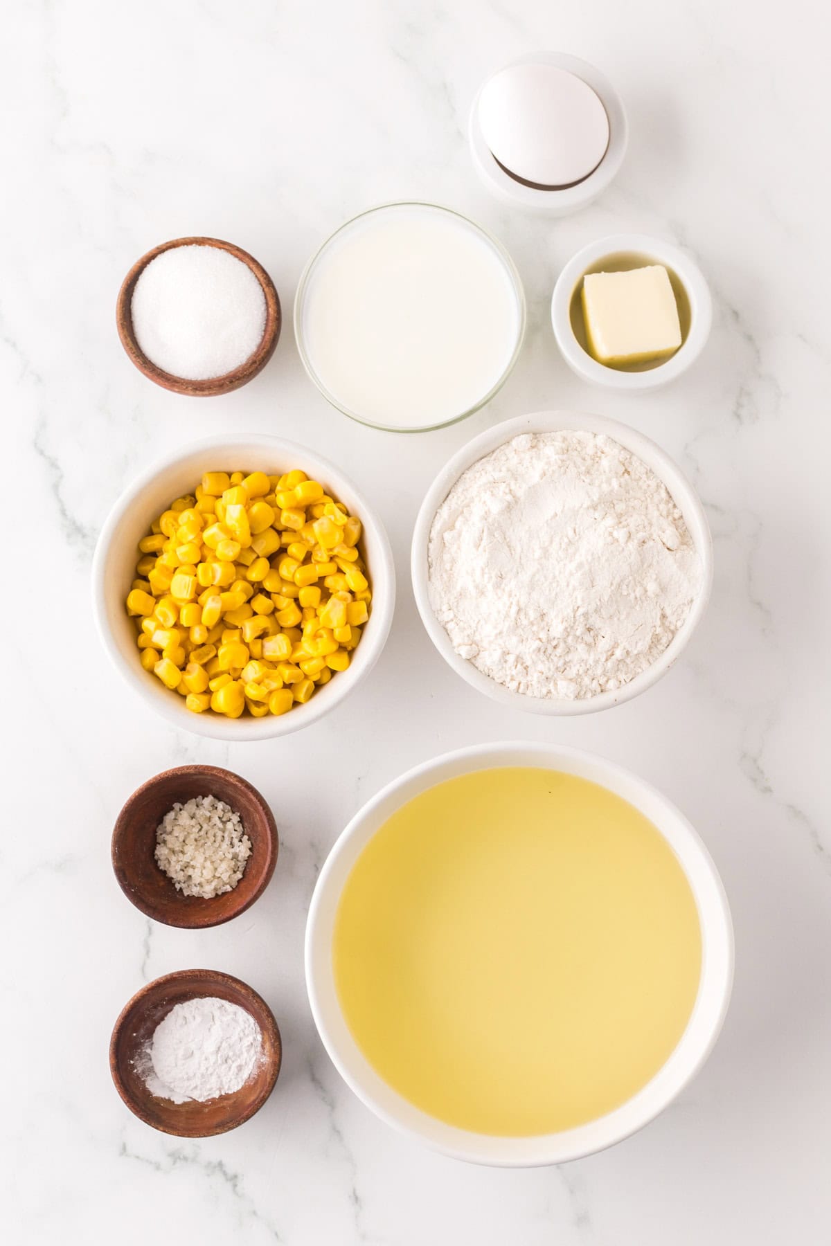 Various baking ingredients are arranged on a white marble surface, including eggs, butter, flour, sugar, milk, corn, and vegetable oil. Small bowls hold salt and baking powder.