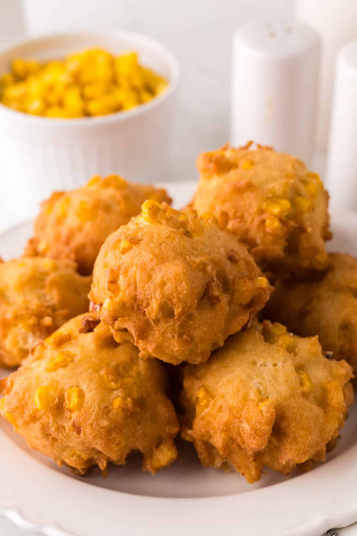 A plate of golden-brown corn fritters stacked in a pyramid shape, with visible corn kernels embedded in the crispy exterior. Behind the plate, a small white bowl filled with corn is partially visible.