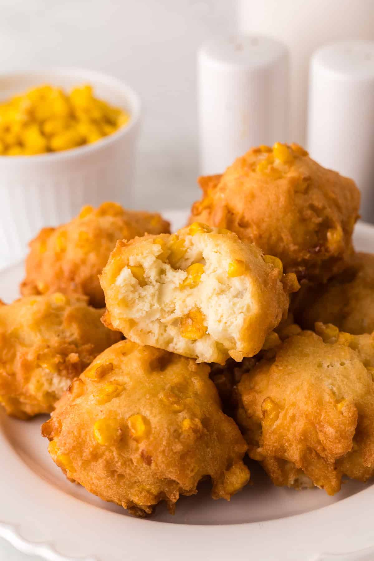 A plate of golden brown corn fritters, one with a bite taken out, revealing the soft interior. A bowl of corn kernels and two salt shakers are in the blurred background.