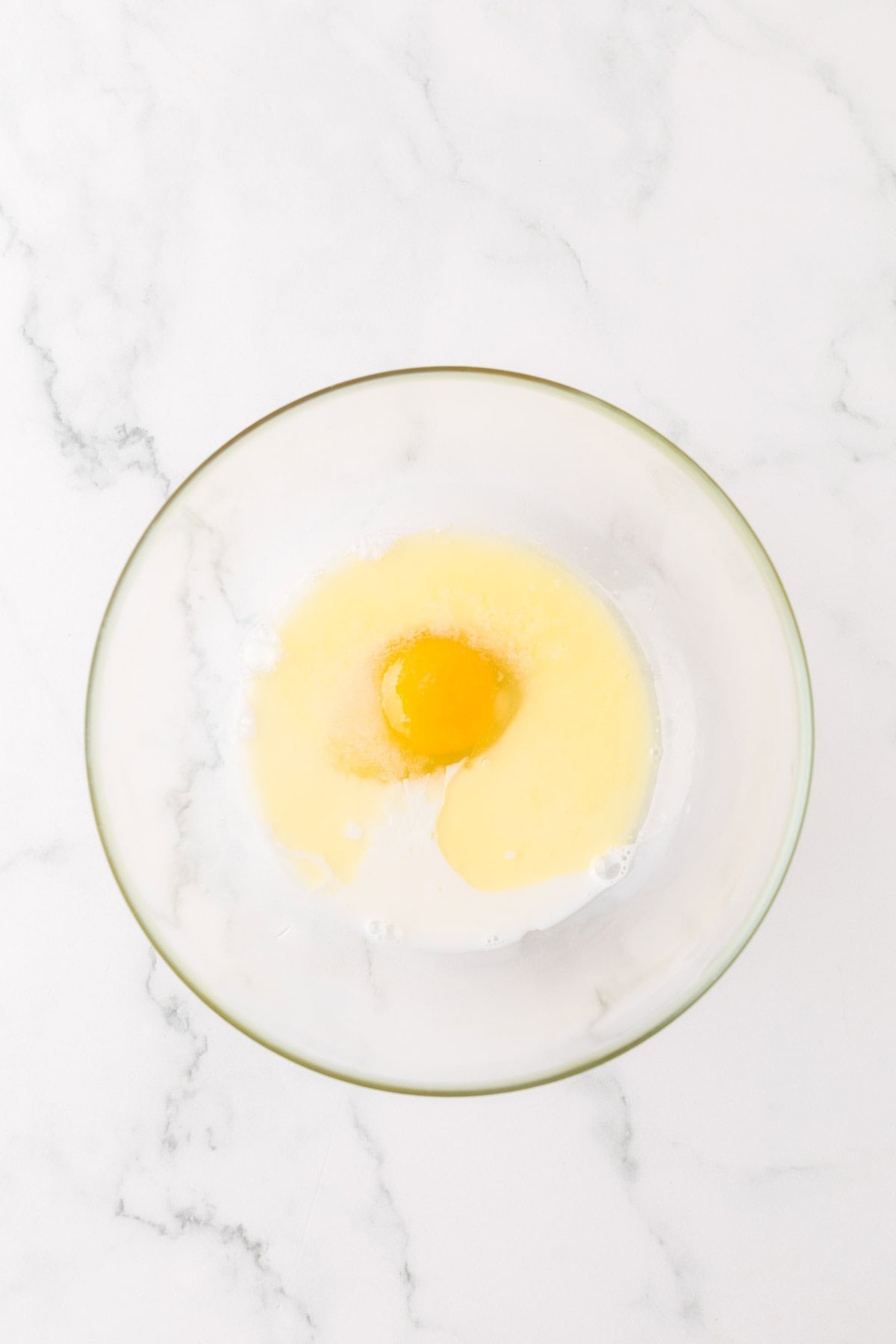 A clear glass bowl on a marble surface contains an egg yolk surrounded by a mixture of melted butter and milk.