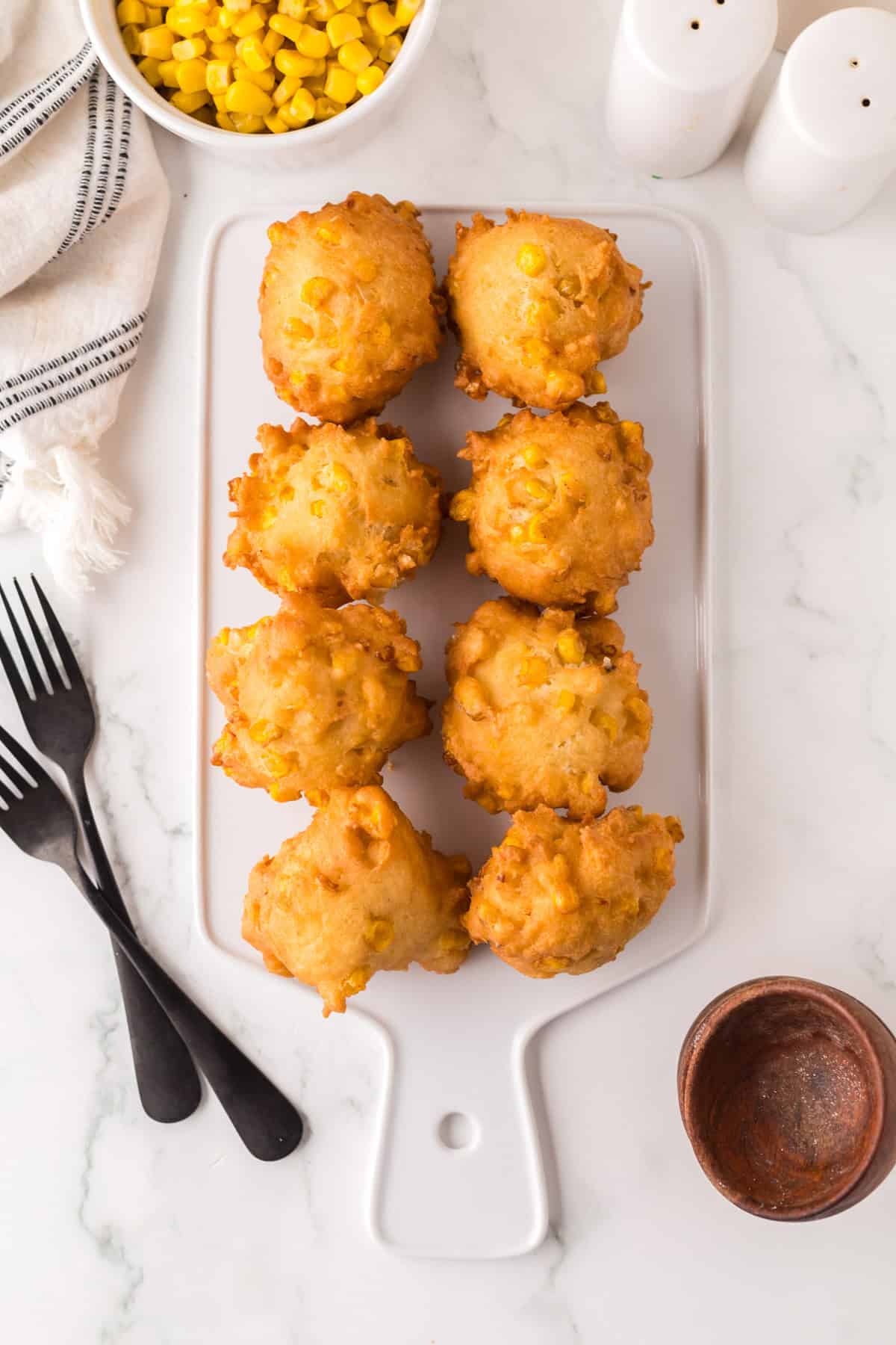 Seven corn fritters arranged on a white rectangular serving platter. Nearby are a small bowl of corn, two black forks, and a wooden dipping dish on a white marble surface.