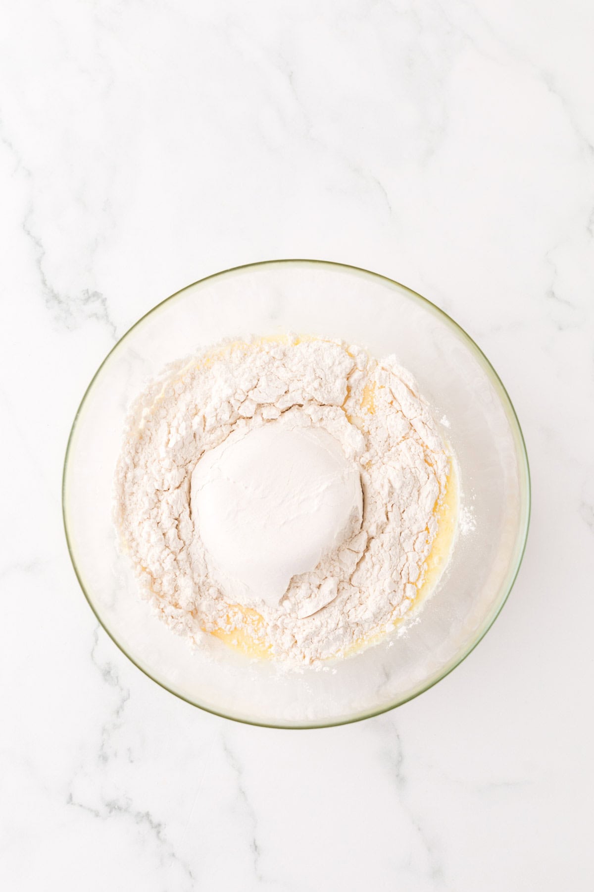 A glass bowl on a marble countertop contains flour and a mound of dough nestled on top of a liquid mixture, possibly butter. The dough is ready for mixing or kneading.