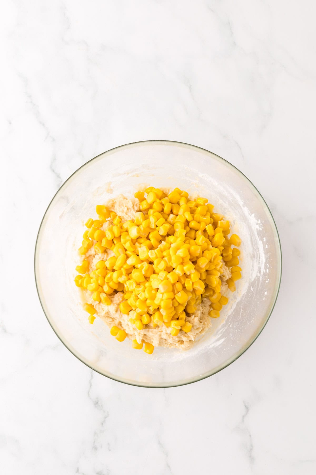 A glass bowl on a marble surface contains a mixture of dough and yellow corn kernels, ready for mixing or baking.