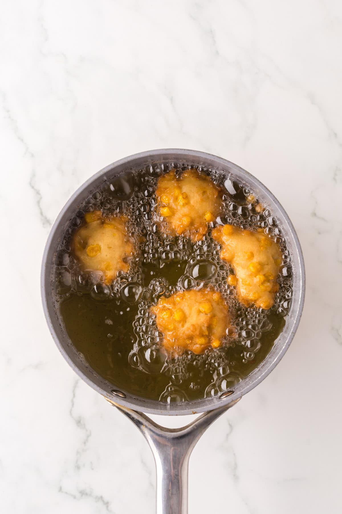 A top view of a small silver-colored skillet with four golden-brown fritters frying in bubbling oil on a marble countertop.