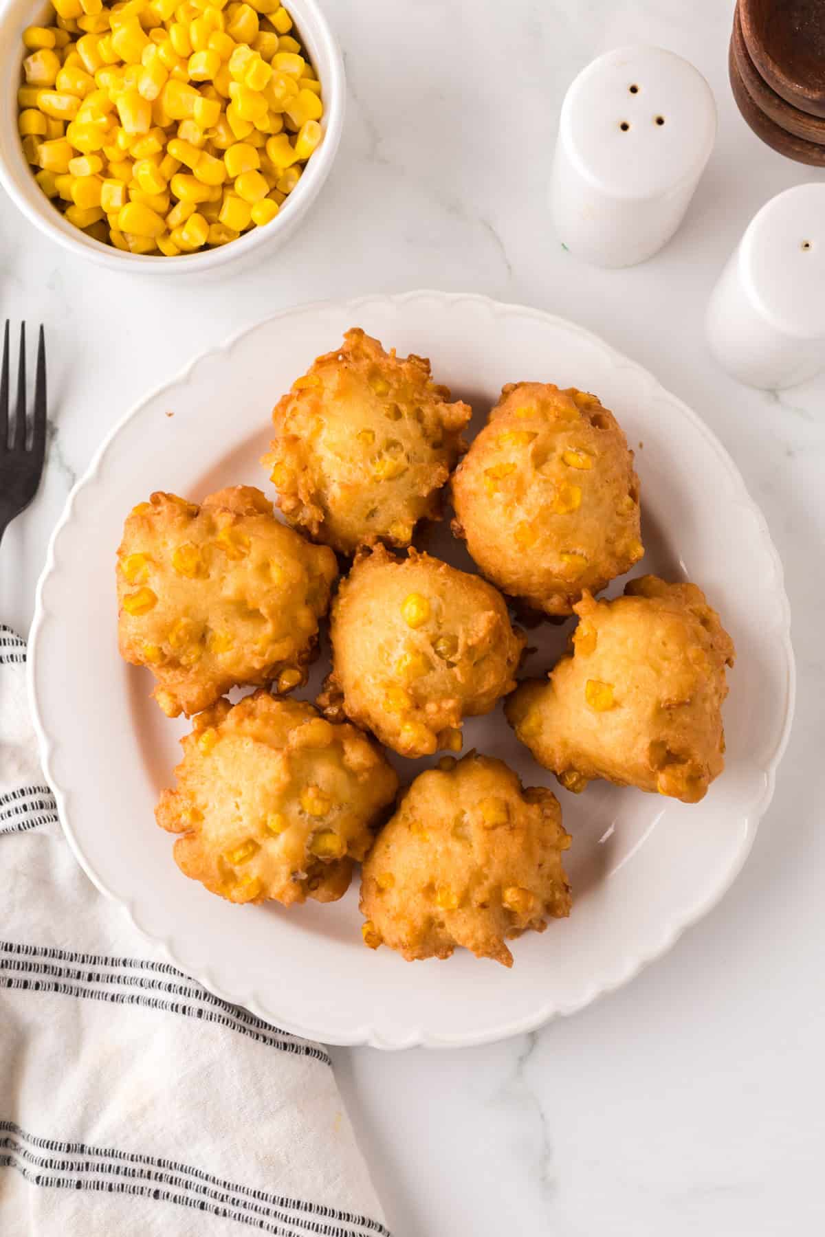 A white plate with seven golden-brown corn fritters, set on a marble countertop. In the background, theres a small bowl of corn kernels and a pair of salt and pepper shakers. A striped cloth napkin and a fork are partially visible.