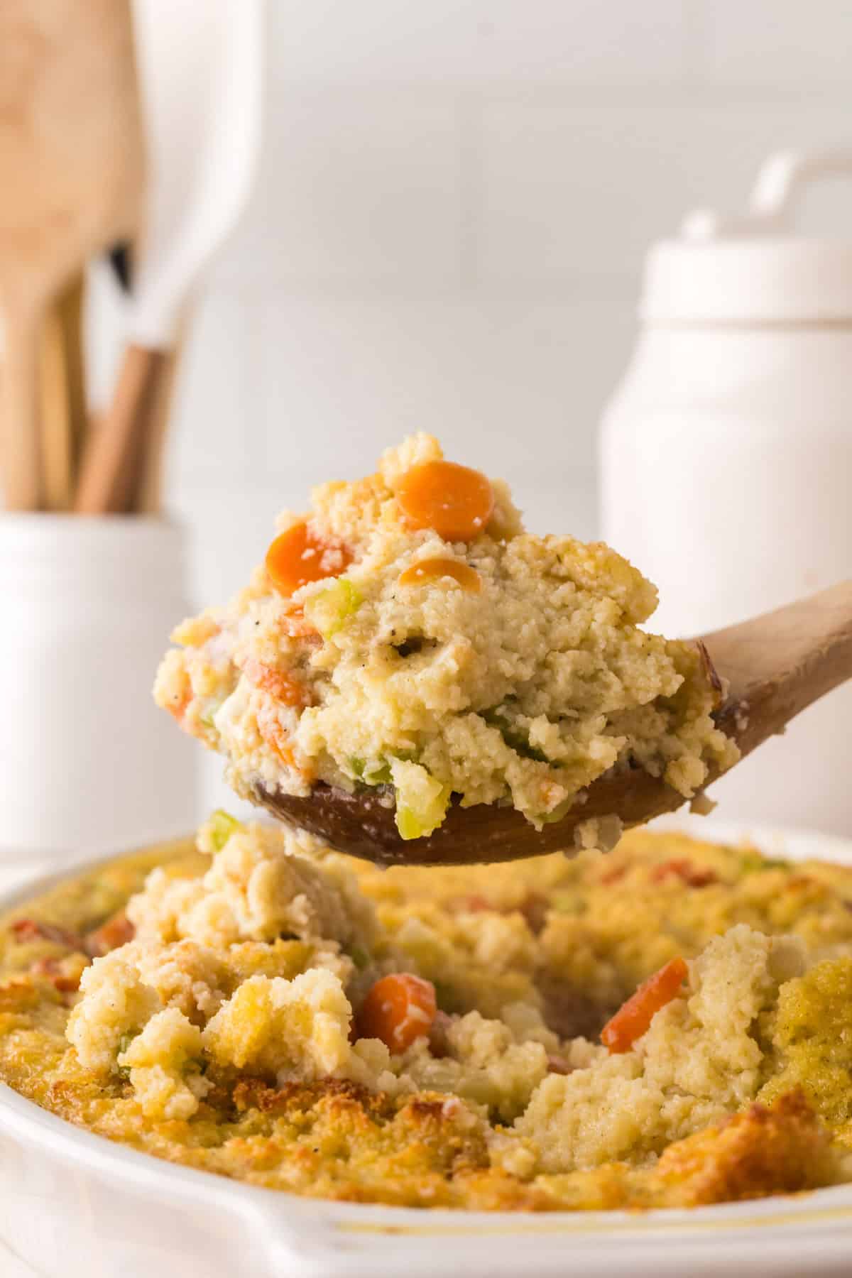 A wooden spoon holds a portion of vegetable cornbread dressing casserole above a white dish filled with the same delight. The casserole showcases pieces of carrots and celery beneath its golden crust. In the background, kitchen containers quietly observe this savory masterpiece.