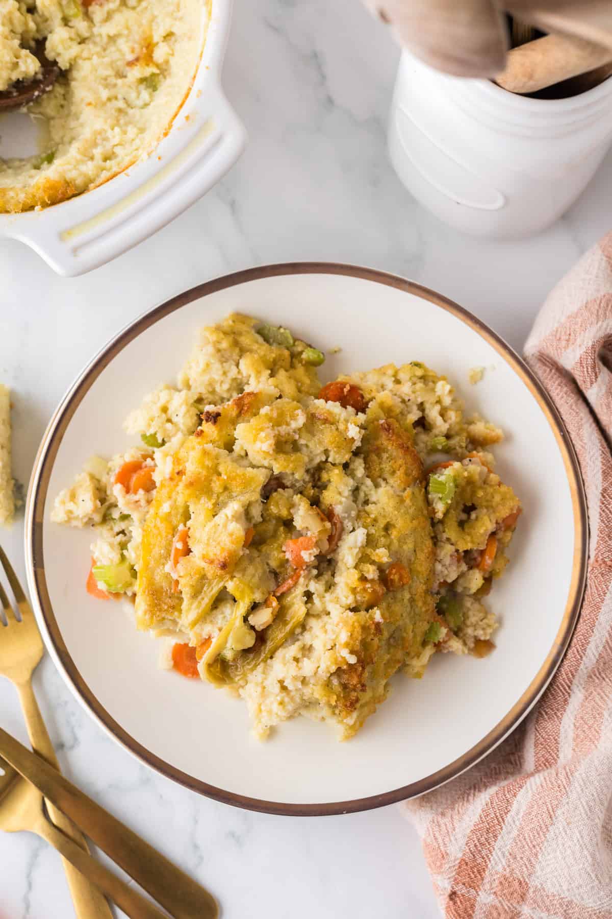 A plate of casserole with vegetables like carrots and peas, complemented by cornbread dressing, served on a white dish with a brown rim. Nearby are a jar with wooden utensils, a gold fork and knife, and a striped cloth. Part of the casserole is visible in a dish in the upper left corner.