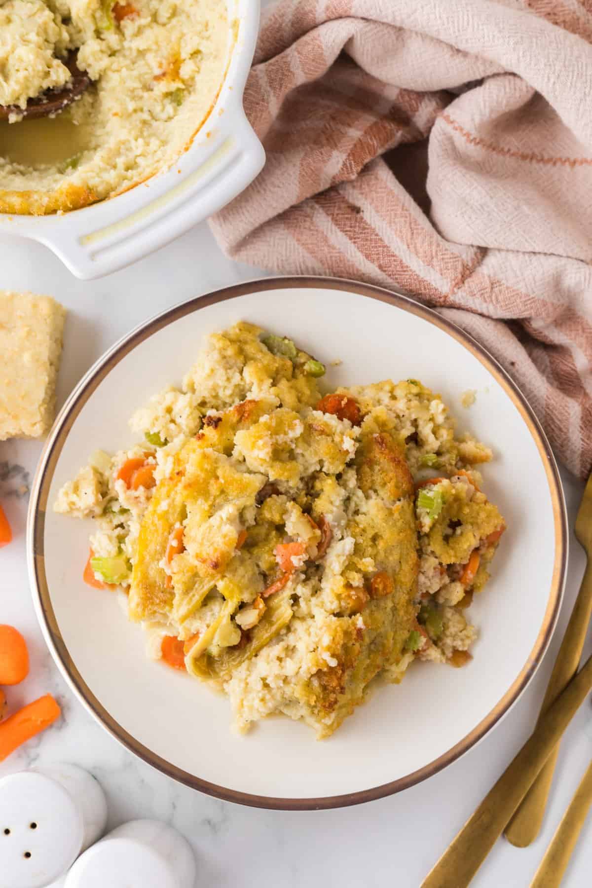 A plate of savory vegetable casserole topped with a golden cornbread dressing, featuring carrots and green vegetables. A portion has been served from a larger casserole dish nearby, with a towel and gold utensils visible in the background.