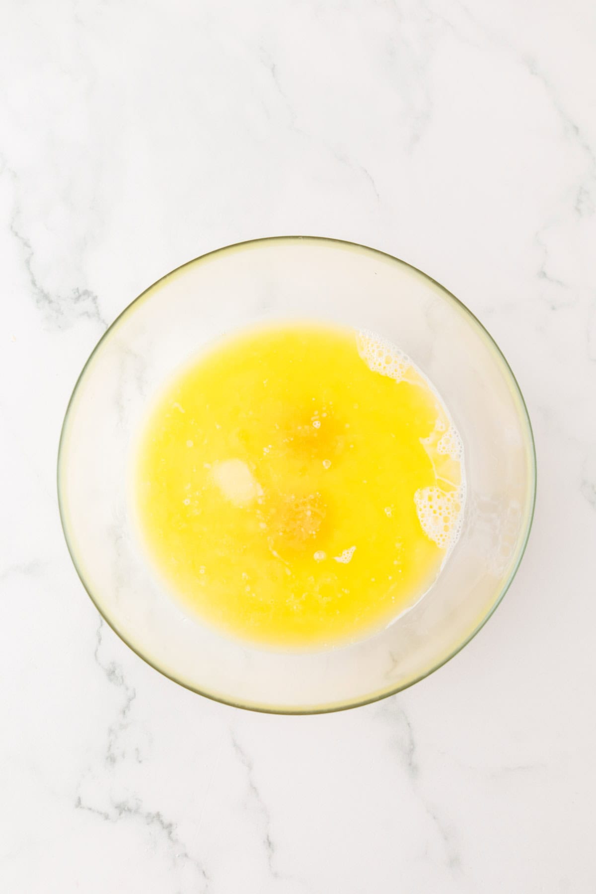 A glass bowl with a yellow liquid mixture, possibly melted butter, sugar, and eggs for cornbread dressing, is on a white marble countertop. The mixture appears slightly frothy.