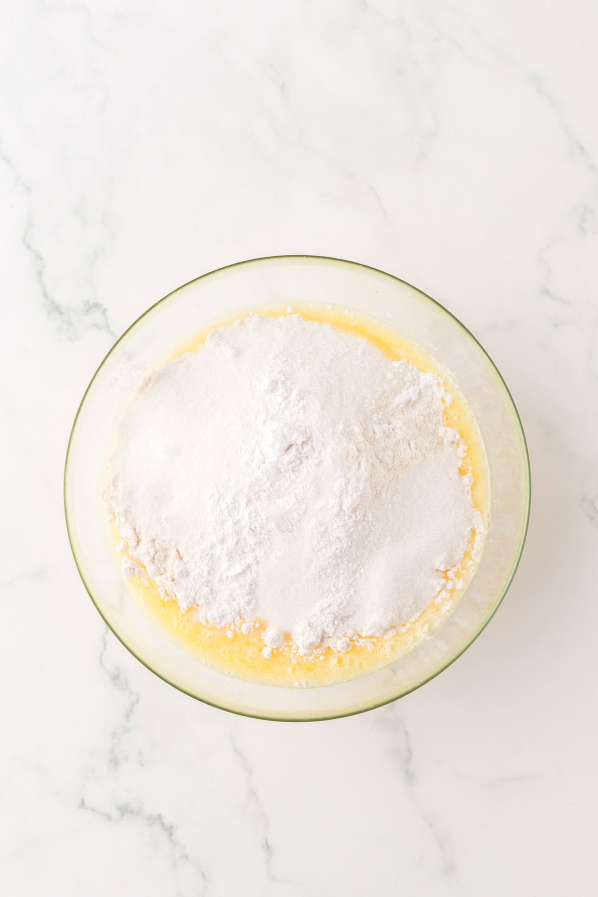 A clear glass bowl containing a mixture of flour and sugar for cornbread dressing sits atop beaten eggs on a marble countertop. The ingredients are unmixed, presenting a striking contrast between the white powder and yellow liquid.