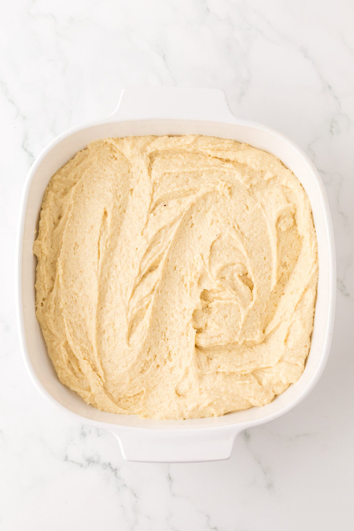 A square white baking dish filled with smooth, swirled cake batter sits invitingly on the marble countertop, ready to be transformed into a delightful creation akin to cornbread dressing.