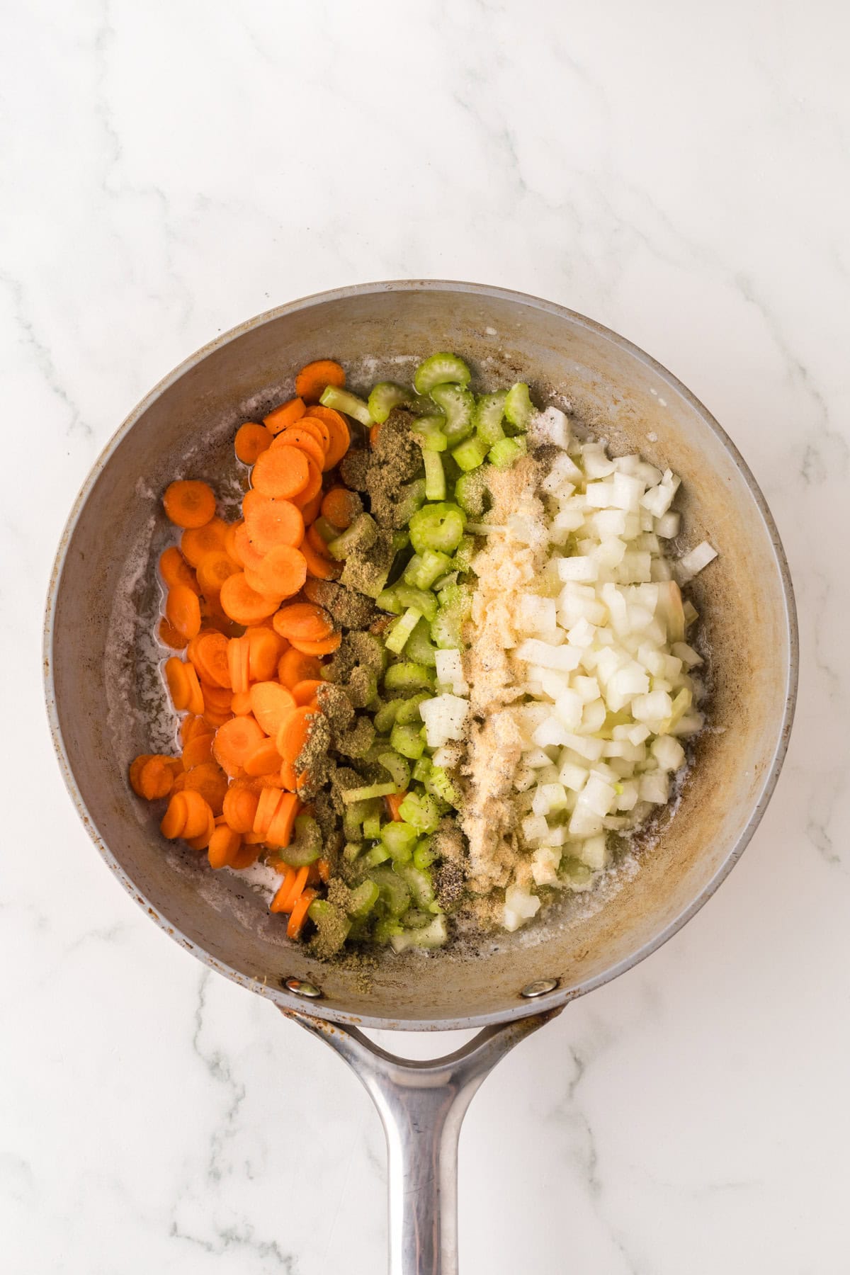 A frying pan on a marble surface contains chopped carrots, celery, and onions being sautéed in butter with a sprinkle of seasoning, forming the flavorful base for comforting cornbread dressing.