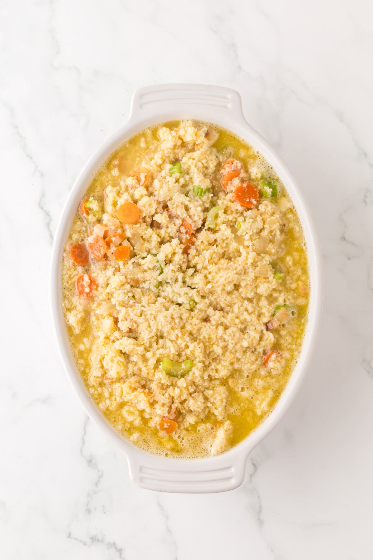 A white oval dish filled with a hearty casserole that combines quinoa, chopped carrots, celery, and colorful vegetables on a white marble surface. The creamy blend is reminiscent of cornbread dressing, making for a warm and comforting meal.