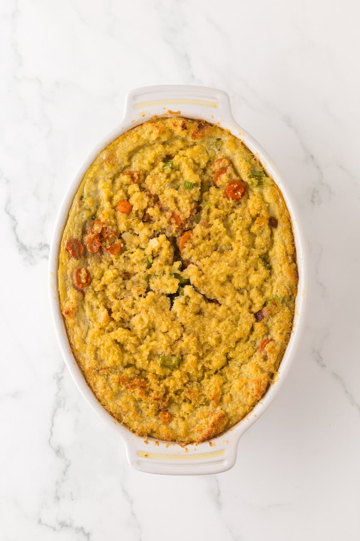 An oval baking dish filled with a golden-brown vegetable casserole, topped with cornbread dressing, features visible pieces of carrots, peas, and other mixed vegetables on a light marble countertop.