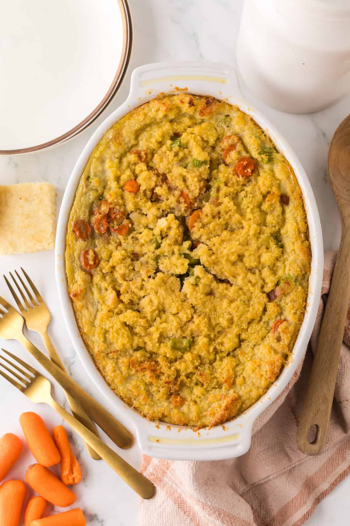 A baked vegetable casserole with a hint of cornbread dressing in a white dish sits on a marble surface. Surrounding the dish are a wooden spoon, gold forks, a stack of plates, and some sliced carrots. The casserole boasts a golden, crispy topping with visible bits of vegetables.