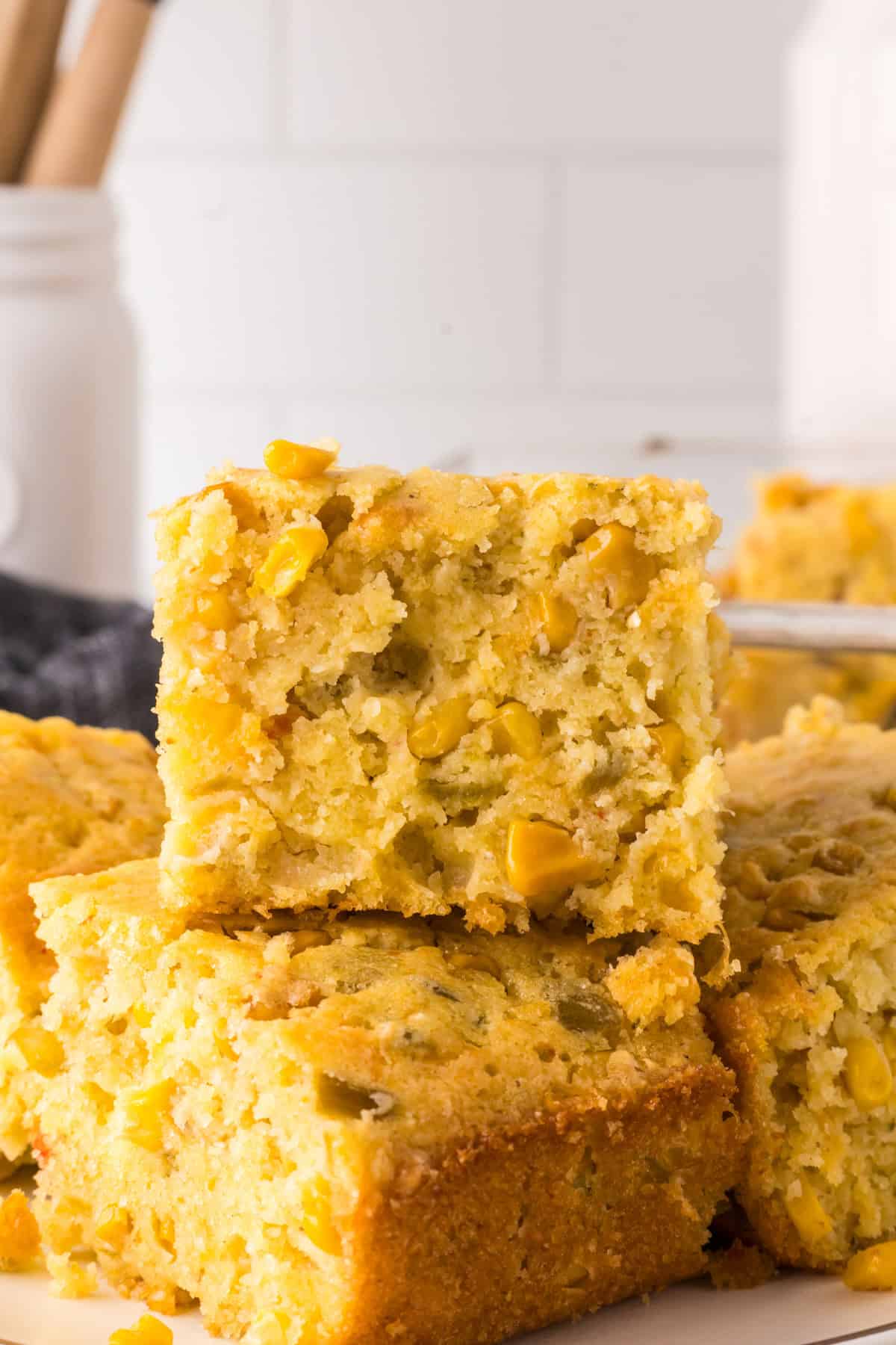 A stack of homemade cornbread slices, featuring visible corn kernels and a golden crust. The background shows a white tiled wall and a blurred jar with utensils.