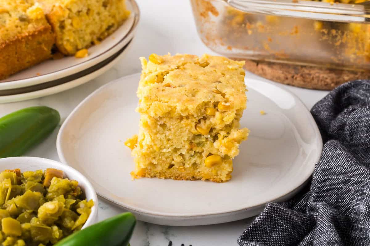 A slice of cornbread with corn kernels sits on a white plate, surrounded by green chili peppers, a bowl of chopped green chiles, and a glass baking dish with more cornbread. A gray napkin is nearby.