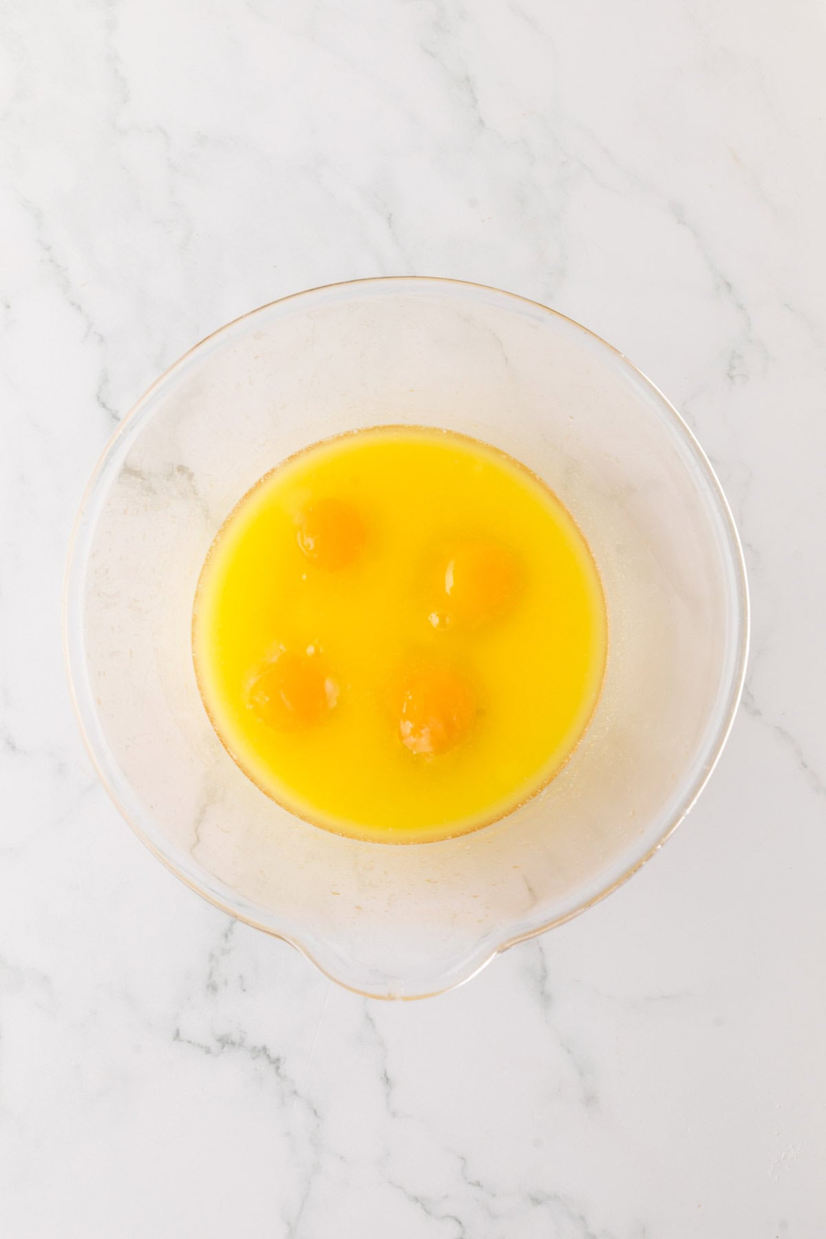 A clear glass bowl with four cracked eggs, showing their bright yellow yolks and whites, sits on a marble countertop.