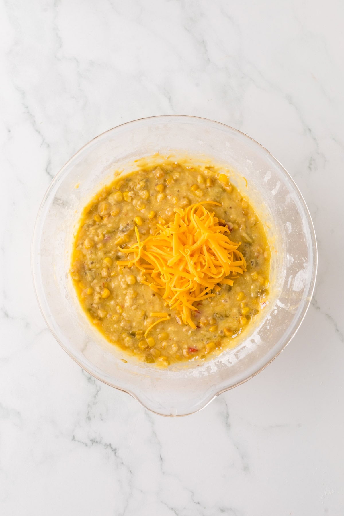 A glass bowl on a marble countertop containing a yellow batter mixed with corn kernels and topped with a small pile of shredded cheddar cheese.