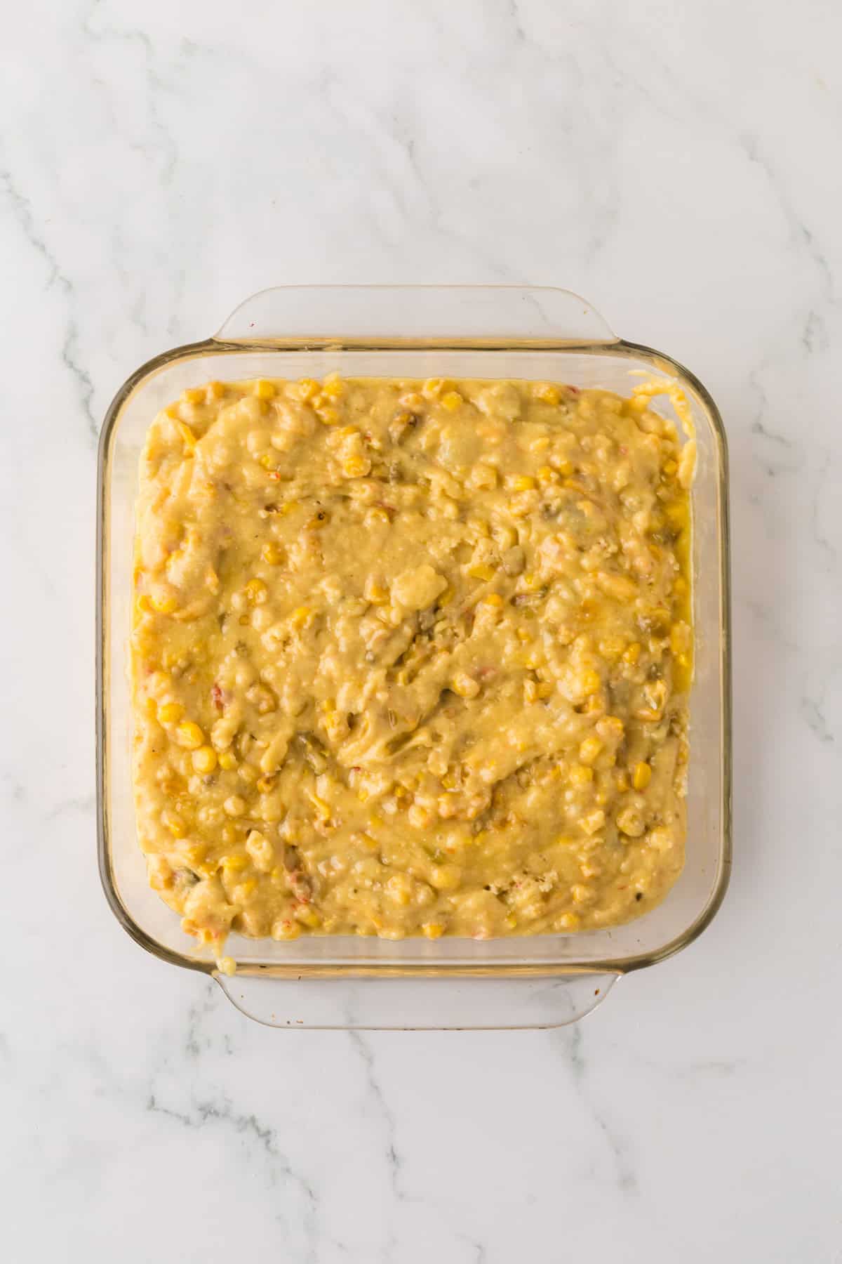 A square glass baking dish filled with a creamy mixture, including visible corn kernels and bits of vegetables, rests on a white marble surface. The dish appears prepped for baking.