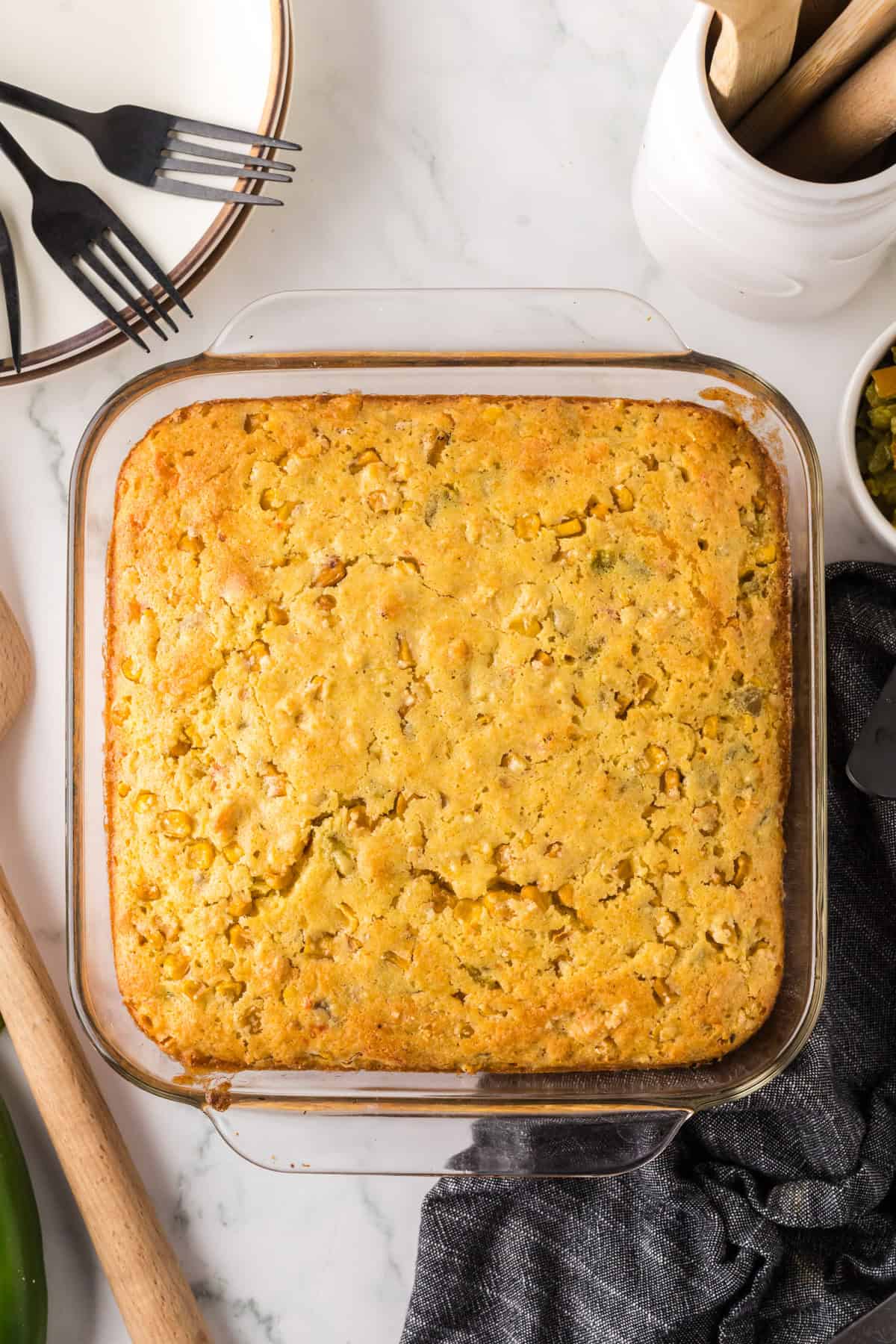 A freshly baked cornbread in a square glass dish on a marble countertop. Nearby are black forks, a white bowl, and a wooden spoon. The cornbread is golden brown, indicating its perfectly cooked.