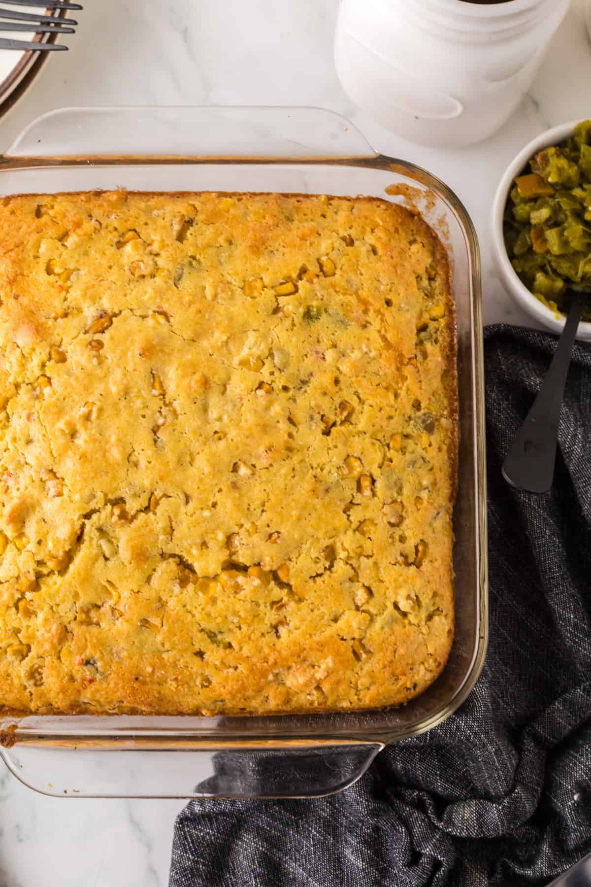 A golden-brown cornbread with chopped green chilies in a square glass baking dish on a marble countertop. A bowl of extra chilies and a dark gray cloth are beside it.