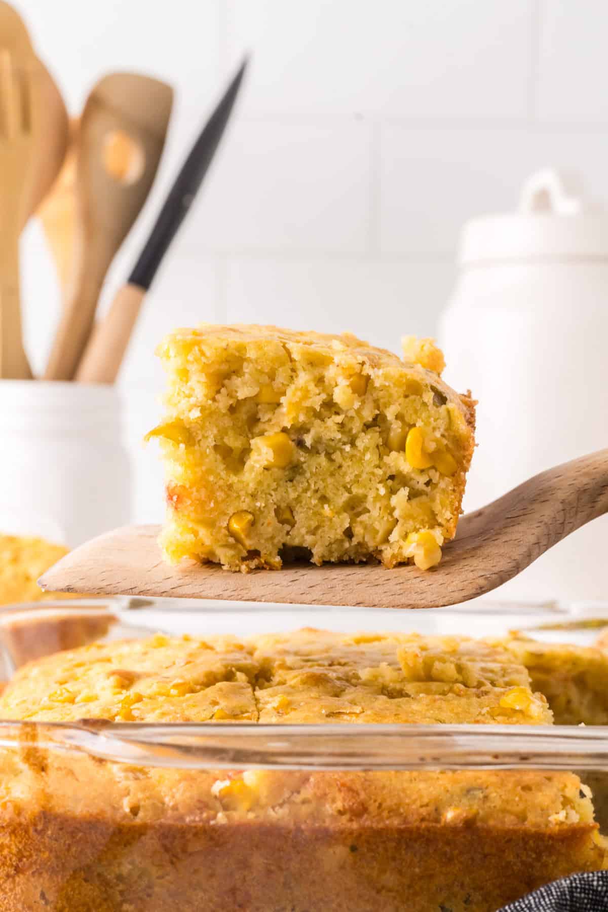 Close-up of a slice of corn casserole on a wooden spatula. The slice is held above a glass baking dish with the remaining casserole. Background includes kitchen utensils and containers against a white tiled wall. The casserole has corn kernels visible.