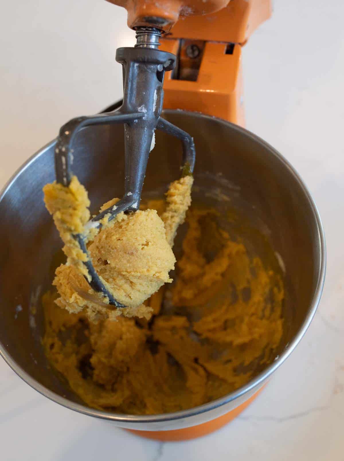 A mixing bowl attached to an orange stand mixer with cookie dough on the beater. The dough is light brown and appears thick, sticking to the metal beater and inside surface of the bowl.