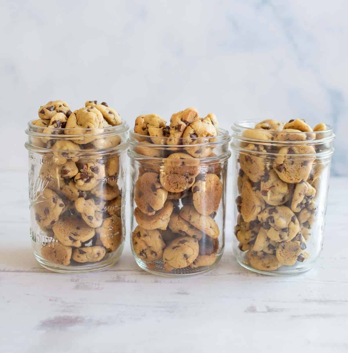 Three glass jars filled with small chocolate chip cookies are lined up on a light wooden surface. The background is a soft, blurred pastel pattern.