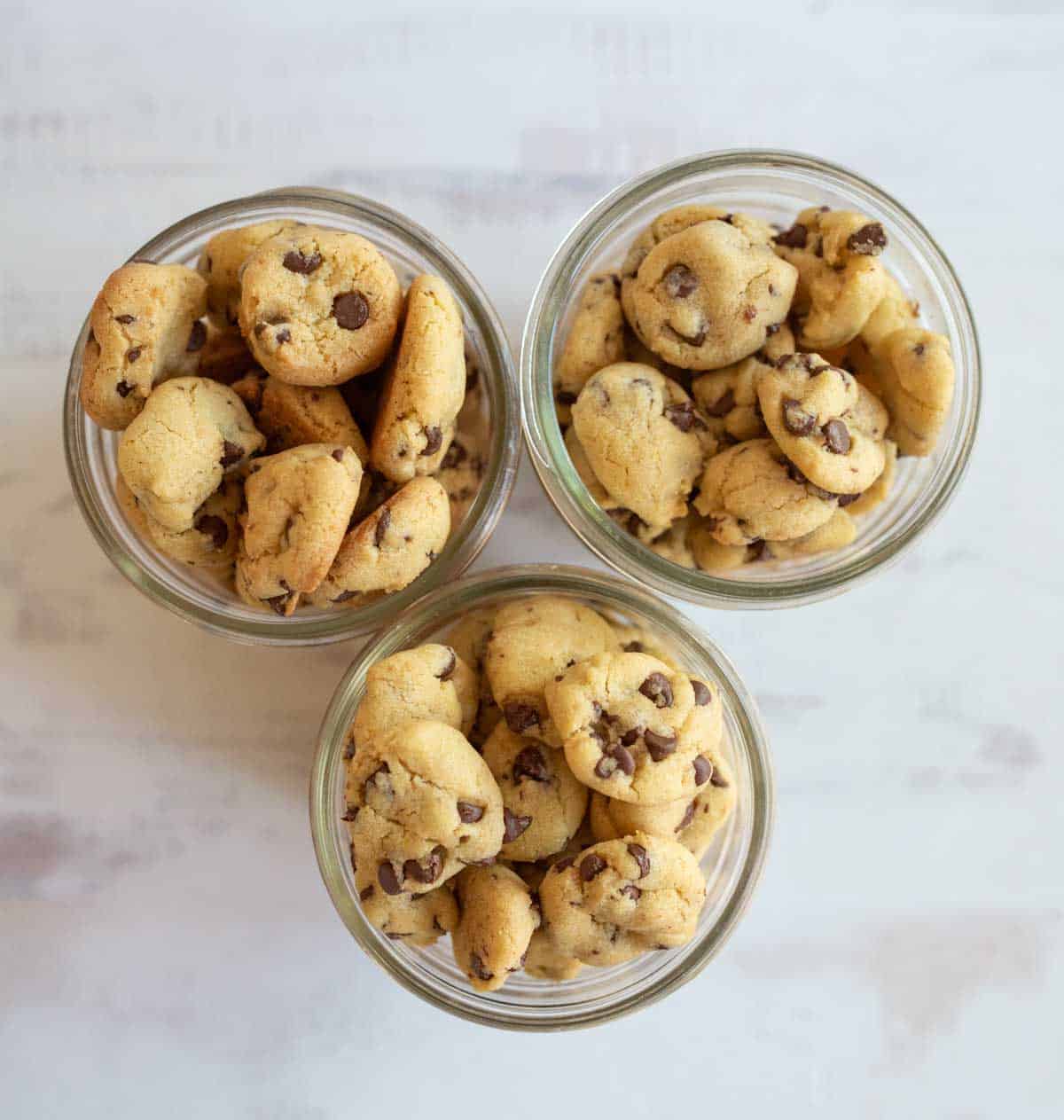 Three glass jars filled with small chocolate chip cookies sit on a light-colored surface. The cookies are stacked neatly, showing their round shape and chocolate chips throughout. The jars are arranged in a triangular formation.