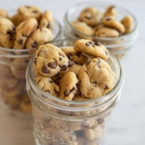 Three glass jars filled with small chocolate chip cookies are placed on a white marble surface. The cookies are golden-brown and plentiful, with visible chocolate chips, creating a tempting display.