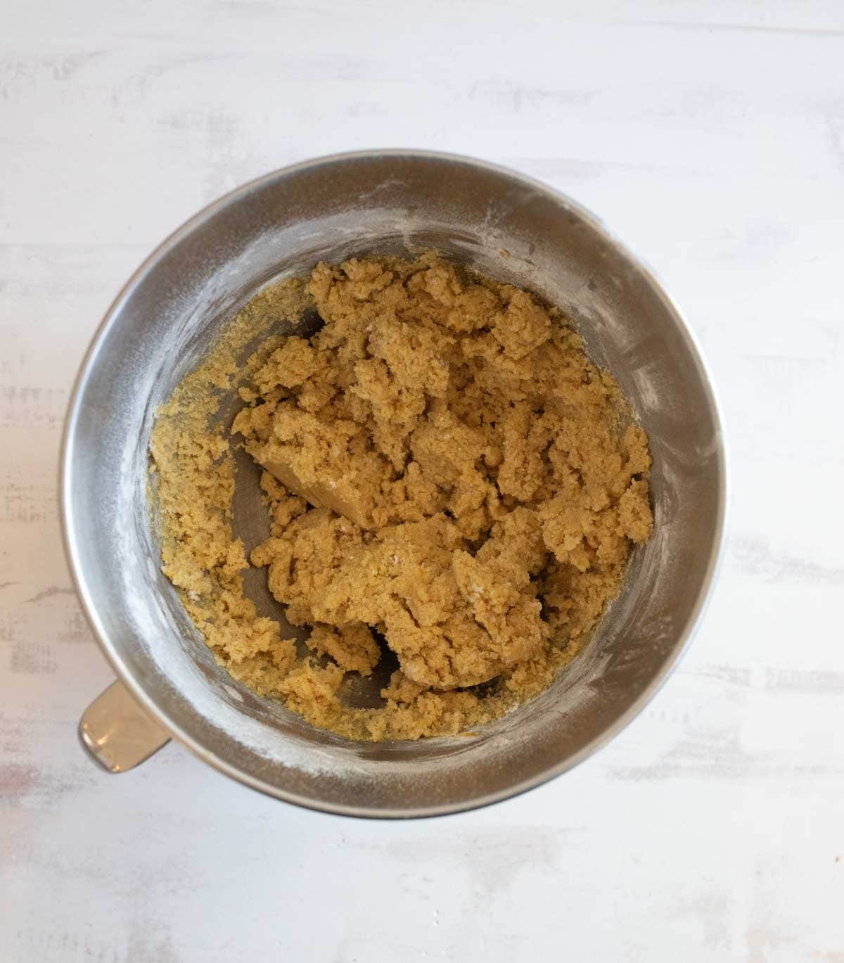 A stainless steel mixing bowl containing a thick, light brown cookie dough on a white surface. The dough is partially mixed, with bits of flour still visible on the sides.