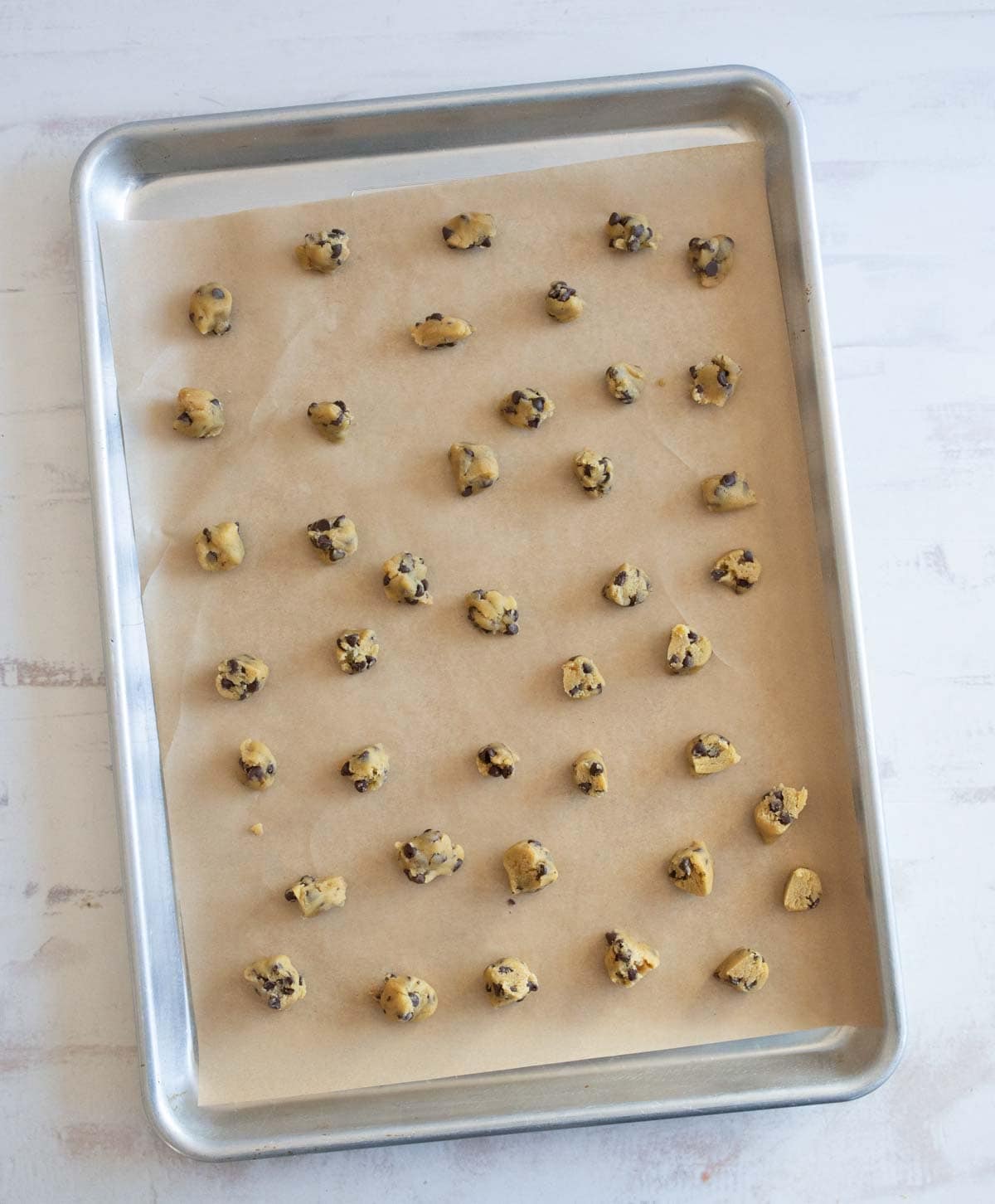 A baking sheet with 36 evenly spaced scoops of chocolate chip cookie dough on parchment paper, ready to be baked. The sheet is placed on a light-colored surface.