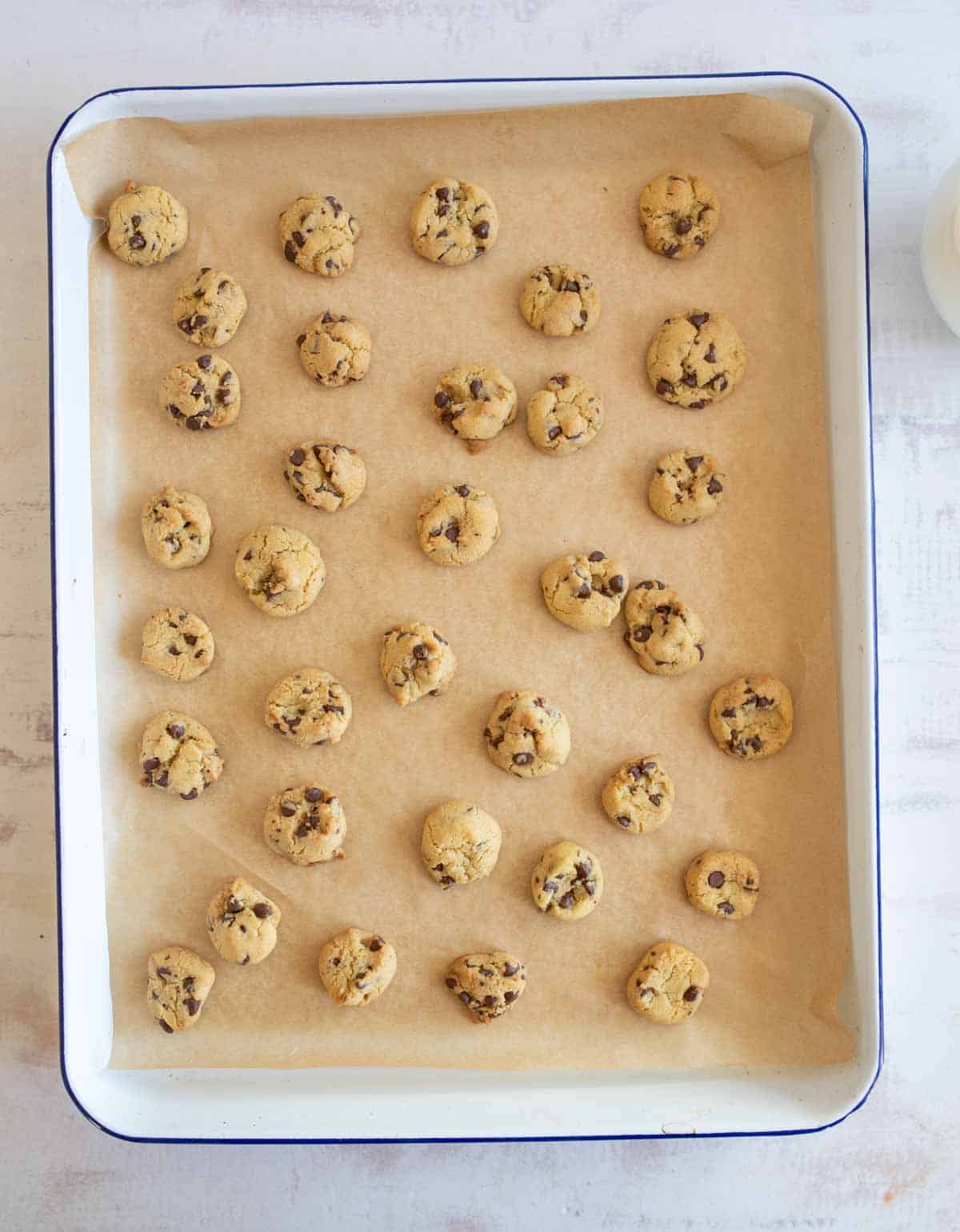 A baking tray lined with parchment paper holds evenly spaced, small chocolate chip cookies. The tray is placed on a light surface, and a bottle of milk is partially visible in the top right corner.