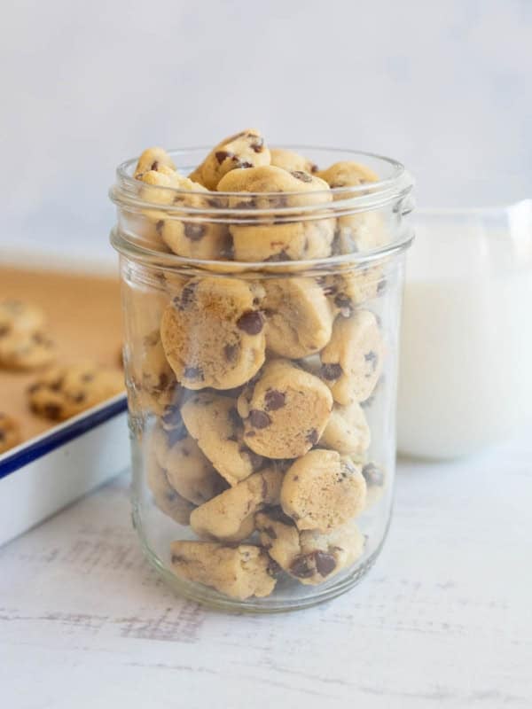 A jar brimming with delicious mini chocolate chip cookies rests on a white surface. Behind it, a glass of milk and a baking tray piled high with more cookies complete the delightful scene, set against a soft, light background.