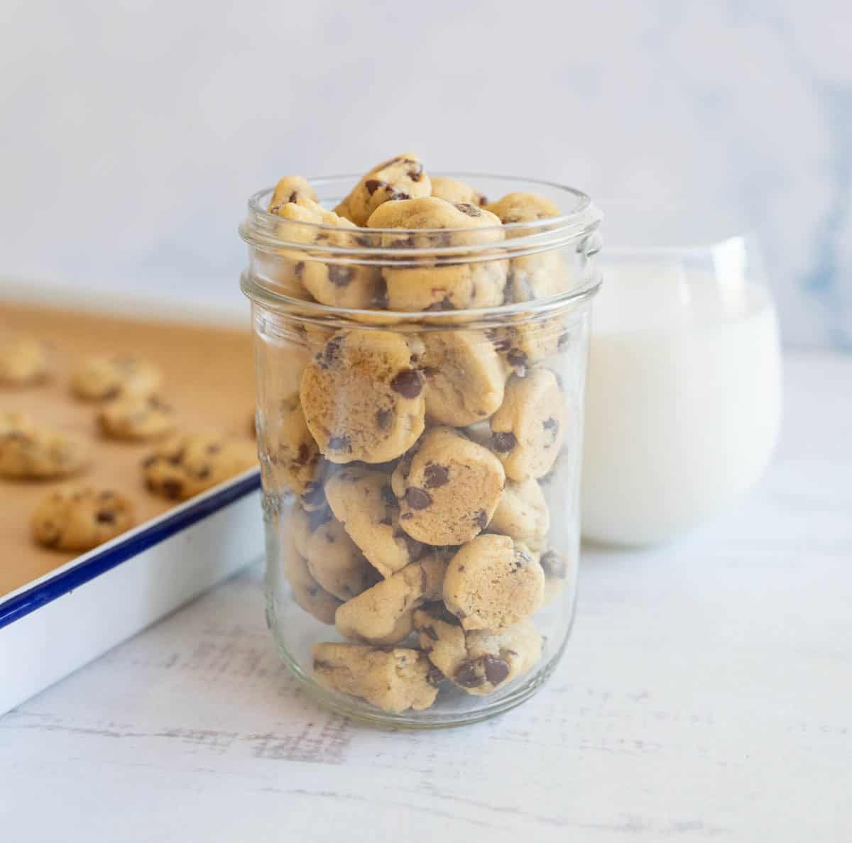 A glass jar filled with mini chocolate chip cookies is placed on a light surface. A glass of milk sits nearby, and a baking tray with more cookies is in the background.