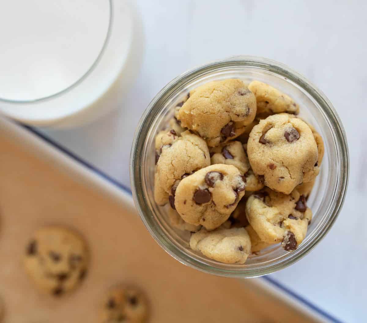 A jar filled with small chocolate chip cookies on a table next to a glass of milk. A baking tray with more cookies is partially visible in the background.