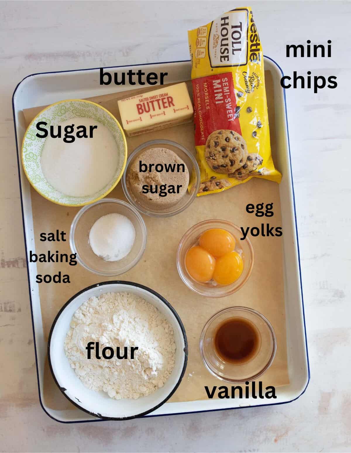 Baking ingredients arranged in a tray: a stick of butter, a bag of mini chocolate chips, sugar, brown sugar, three egg yolks, vanilla extract, flour, salt, and baking soda. Each item is labeled.