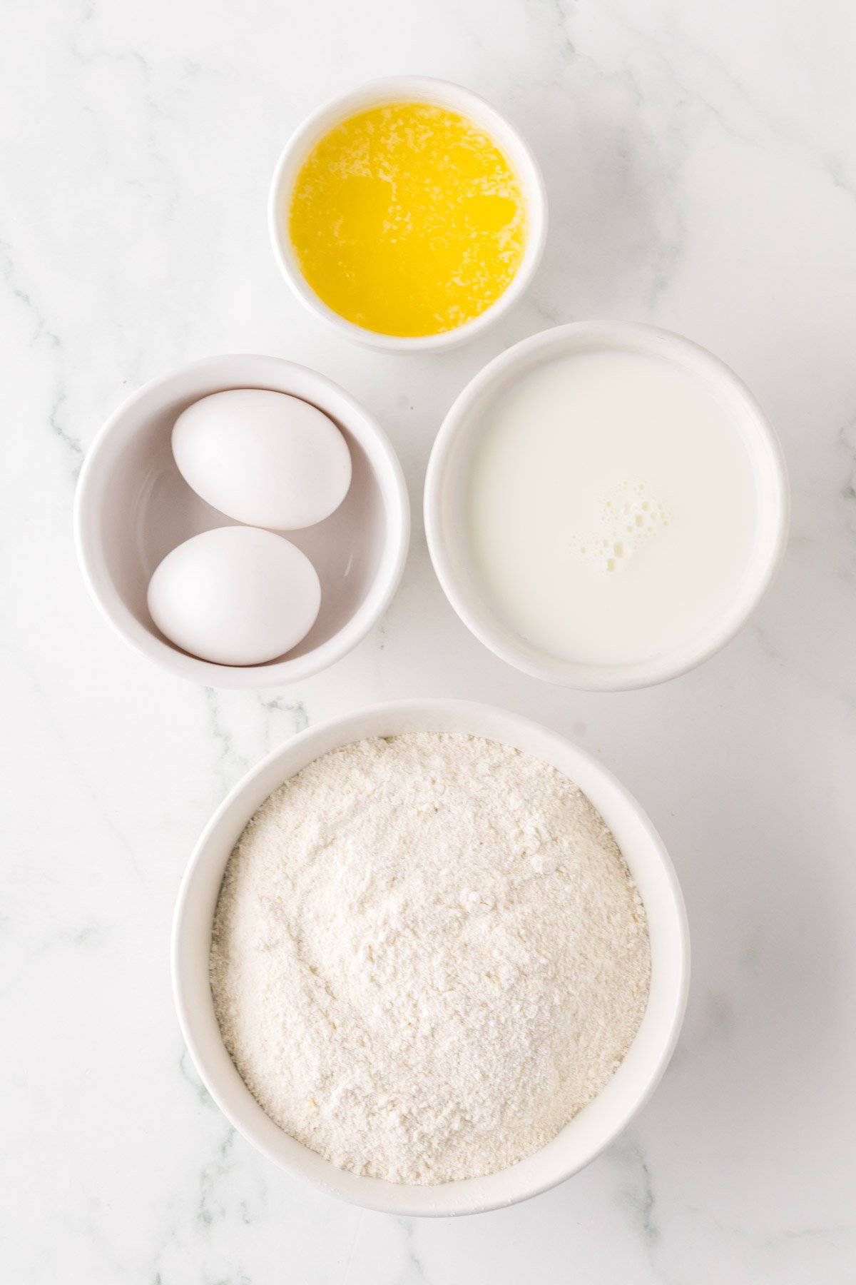 A top-down view of baking ingredients on a marble surface. Includes a bowl of flour, two eggs in a separate bowl, a small bowl of melted butter, and a bowl of milk, all arranged neatly.