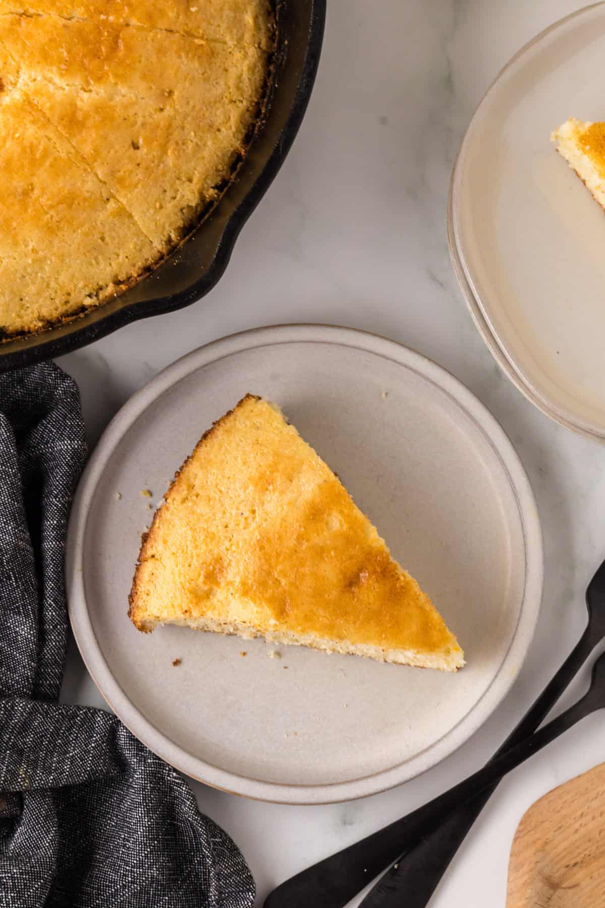 A slice of cornbread is served on a round, light gray plate. The whole cornbread, partially visible in a pan, is in the top left corner. A dark cloth napkin is nearby on a white marble surface.