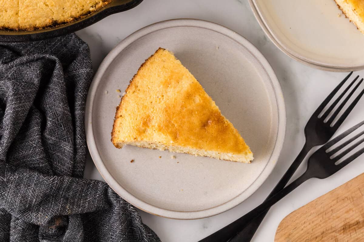 A slice of cornbread on a round, light gray plate. The cornbread has a golden-brown crust. Two black forks lie nearby on a table with a dark napkin. Another plate with a partial view of more cornbread is in the background.